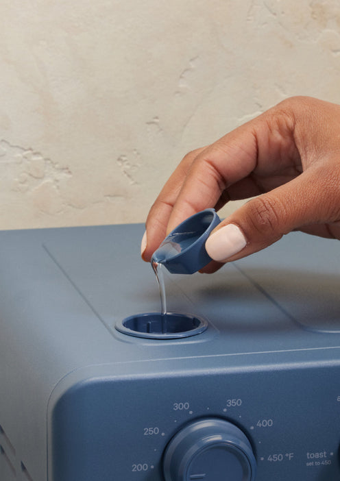 A hand pours liquid from a small cup into the top opening of a blue kitchen appliance with a temperature dial.
