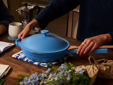 Person holding a blue casserole dish on a wooden table with flowers and a coffee maker in the background.