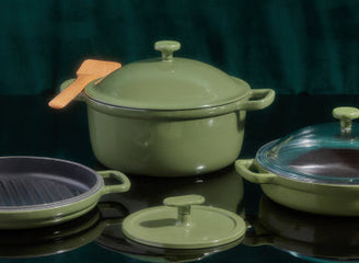 Three green kitchen pots and pans with matching lids, one with a wooden spatula resting on top, set against a dark green textured background.