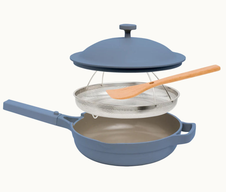 A blue nonstick pan with a matching lid, a stainless steel steamer basket, and a wooden spatula, all shown separated and stacked above the pan.