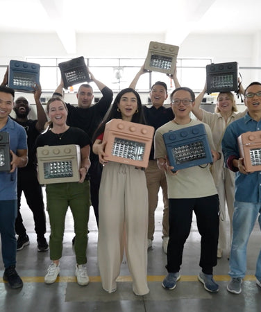 A group of people stand indoors holding portable ovens and smiling towards the camera, with some raising the ovens above their heads.