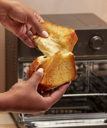 A person tears a piece of golden brioche bread in front of an open countertop oven.