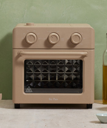 Beige countertop oven with three dials, a matching lidded pan, kitchen utensils in a glass, a bottle of oil, grapes in bowls, and a blue vase are arranged on a light-colored surface.
