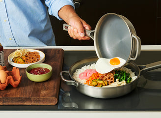 Person cooking an egg over rice with vegetables in a frying pan on a stove.