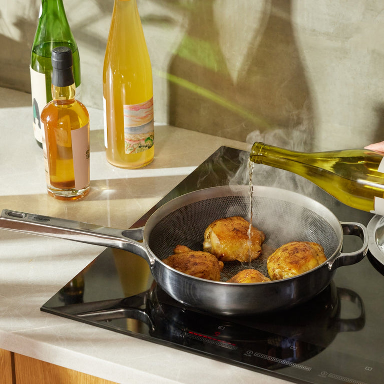 A person pours white wine from a bottle into a frying pan with browned chicken thighs on an induction stove, with bottles on the countertop nearby.