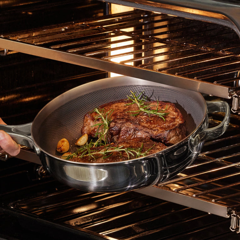 A hand holds a metal pan with cooked steaks, garlic cloves, and herbs, placing it into an open oven with metal racks.