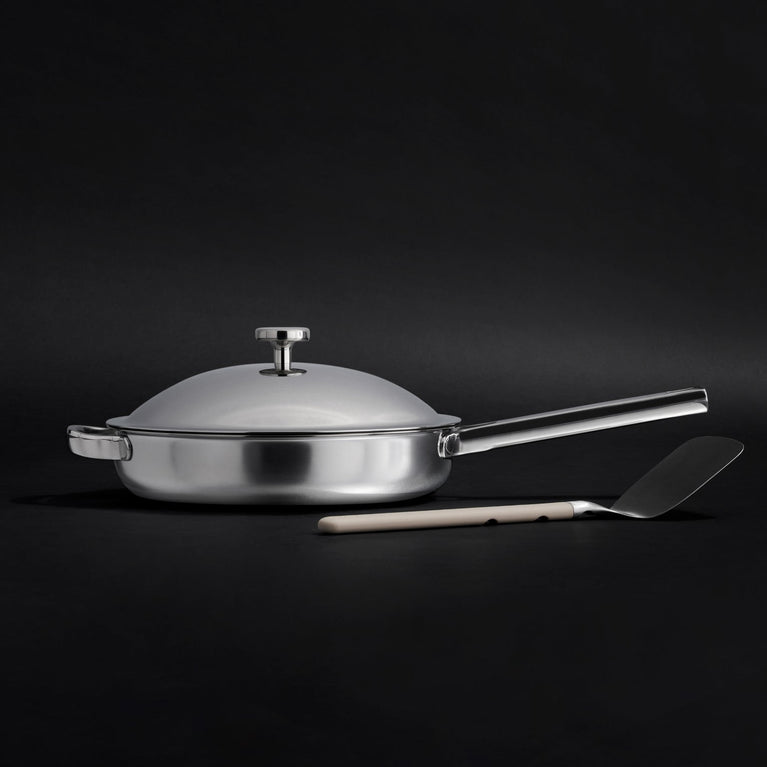 A white frying pan with a lid and a stainless steel handle is placed on a light surface, accompanied by a beige-handled spatula in front.