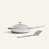 A white frying pan with a lid and a stainless steel handle is placed on a light surface, accompanied by a beige-handled spatula in front.