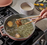 A hand uses chopsticks to deep fry battered vegetables in a pink skillet on a stove, with a bowl of batter and sliced vegetables nearby.