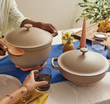Two people serve food at a table set with beige cookware, plates, glasses, a candle, and a small vase of yellow flowers on a blue table runner.