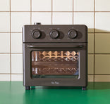 A compact countertop oven with three control knobs sits on a green surface in front of a tiled wall.