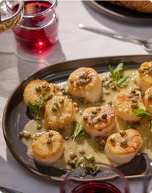 Plated scallops with a creamy sauce and capers on a table with red wine.