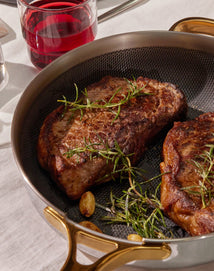 Steaks with herbs in a pan on a table with a glass of red wine.