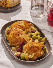 Plated dish of roasted chicken with olives on a table with glasses of red wine.