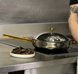A stainless steel pan with a gold handle and lid sits on a stovetop next to a plate of sliced dark food. A persons tattooed arm is partially visible on the right.