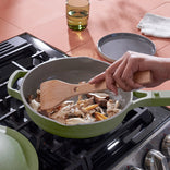 A person stirs assorted mushrooms in a green pan on a gas stove using a wooden spatula; a green glass and plate are in the background.