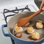 A hand uses a wooden spatula to cook four seared scallops in a blue nonstick skillet on a gas stove.