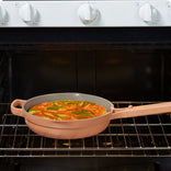 A round frittata with green vegetables is cooking in a pink nonstick skillet on the middle rack of an oven with white control knobs.