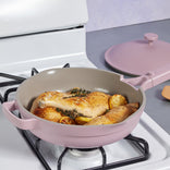 Three seasoned chicken drumsticks with lemon slices and herbs cook in a pink nonstick pan on a stovetop, with the matching pink lid resting nearby.