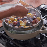 A hand holds a dish towel while lifting a steamer basket of assorted small potatoes from a pot of boiling water on a stovetop.