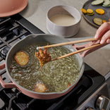 A hand uses chopsticks to fry battered food pieces in hot oil on a stovetop. A bowl of batter and sliced vegetables are in the background.
