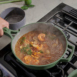 A person cooks shrimp and vegetables in a green pot on a gas stove, with steam rising from the simmering mixture.