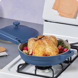 A whole roasted chicken with radishes and herbs in a blue skillet on a stovetop, with the lid resting nearby.