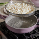 A metal strainer with cooked noodles is held over a pink pot of hot water on a stovetop, allowing the water to drain.