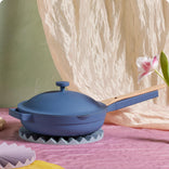 A blue lidded frying pan with a wooden handle sits on a trivet atop a pink surface, with a flower and draped fabric in the background.