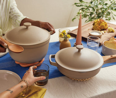 Two people share a meal at a table set with beige cookware, plates, glasses, a candle, and food, including roasted cauliflower on a wooden board.