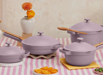 Four lavender-colored cookware pots and pans with wooden handles are arranged on a pink striped tablecloth, with vases and sliced oranges in the background.