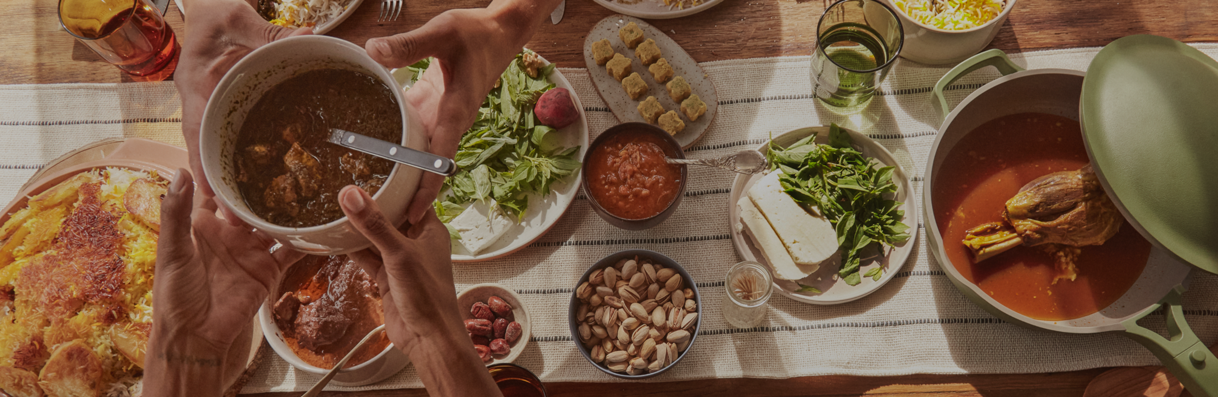 A table set with various dishes, including a pot with meat in broth, salad, bread, nuts, sauces, and hands serving food.