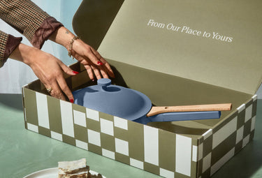 A person places a blue pan with a wooden handle into a patterned gift box labeled From Our Place to Yours.