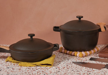 Two black cast iron cookware pieces with lids sit on a countertop; one is on a yellow cloth, the other on a round felt trivet near two kitchen knives.