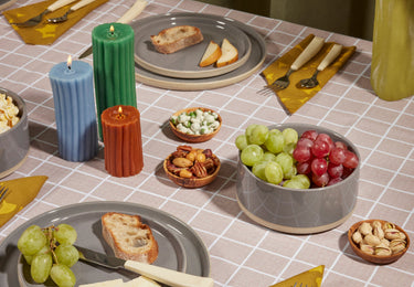 A table set with plates of bread, cheese, grapes, nuts, and snacks, surrounded by yellow napkins, utensils, and three colorful ribbed candles.