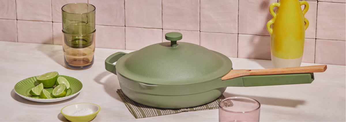 Translation missing: en.A green lidded pan with a wooden handle sits on a countertop near a bowl of lime wedges, glasses, a salt dish, and a yellow vase, against a tiled backsplash