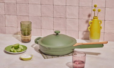 Translation missing: en.A green lidded skillet with a wooden handle sits on a countertop beside stacked glasses, a plate of lime wedges, a small bowl, and a yellow vase with flowers