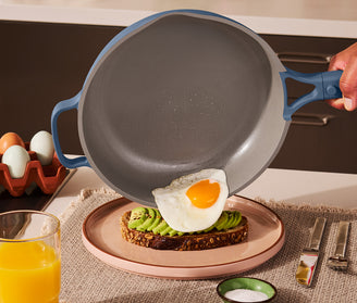 A hand slides a fried egg from a nonstick pan onto avocado toast on a plate. A glass of orange juice, utensils, salt, and eggs in a carton are on the table.