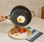 A hand tilts a frying pan to slide a fried egg onto a plate with toast, roasted tomato, a grilled green pepper, and bacon on a kitchen counter.