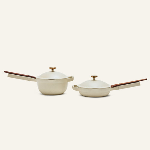 The Mini Cookware Duo features two cream-colored pots with wooden handles and lids, including the versatile Mini Always Pan, displayed side by side to showcase stylish and multifunctional cookware.